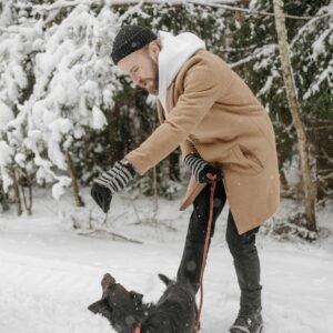 A bearded man in winter clothes plays with his dog on a snowy path in a forest.