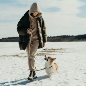 A woman in a puffer jacket walks her corgi on a snowy day, showcasing winter fashion and companionship.