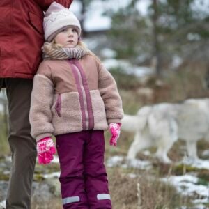 Child in warm clothes standing outdoors, with a snowy background and dog nearby.