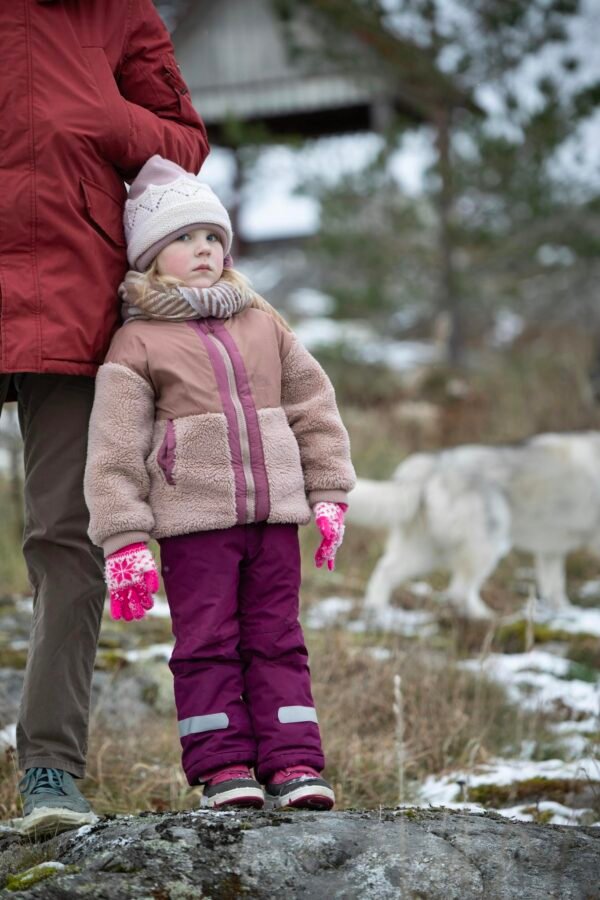 Child in warm clothes standing outdoors, with a snowy background and dog nearby.